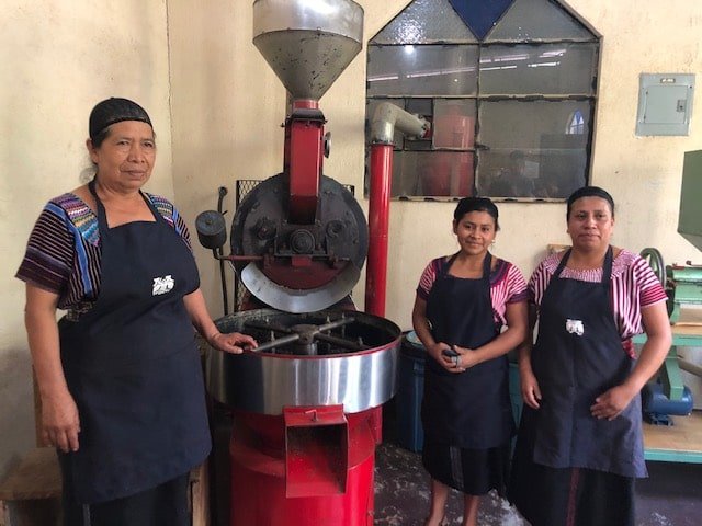 Three people wearing aprons stand beside a red coffee roaster.